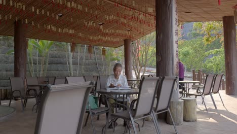 Anciano-Chino-Sentado-En-Una-Zona-De-Asientos-Al-Aire-Libre-Bajo-Un-Techo-Leyendo-Un-Libro-Solo