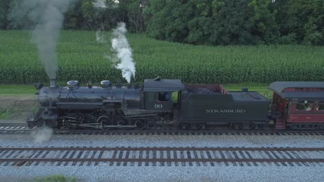 Vista-Aérea-De-Un-Motor-De-Vapor-De-1924-Con-Un-Tren-De-Pasajeros-Que-Viaja-A-Lo-Largo-De-La-Campiña-Amish-Mientras-Las-Puestas-De-Sol-En-Un-Día-De-Verano-Visto-Por-Un-Dron