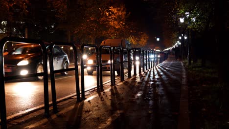 Cars-moving-slowly-on-city-street-at-night,-lights-flare-shining-through-railing-irons-bordering-sidewalk