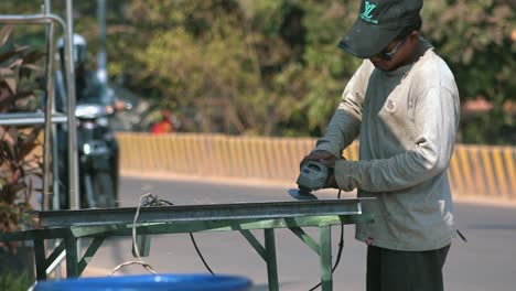 Tiro-Medio-Lento-De-Un-Hombre-Asiático-Al-Lado-De-La-Carretera-Usando-Una-Amoladora-Angular-En-Un-Trozo-De-Metal