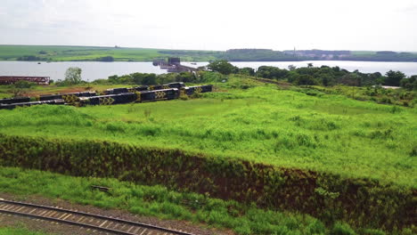 Stationäre-Eisenbahnwaggons,-Die-Darauf-Warten,-Beladen-Zu-Werden,-Wunderschöne-Grüne-Vegetation-Rundherum,-Im-Hintergrund-Ein-Wunderschöner-Fluss,-Eine-Eisenbahnbrücke-Und-An-Der-Küste-Angedockte-Boote