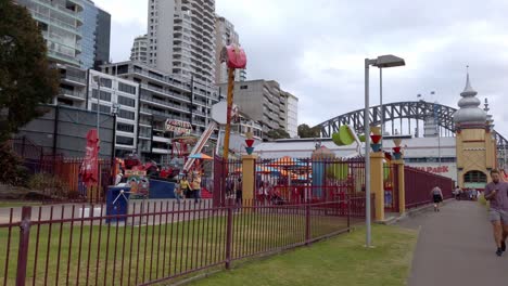 Luna-Park-in-the-waterfront-area-of-northern-shore-on-an-overcast-day-with-Harbour-Bridge-in-the-distance,-Stable-walking-handheld-shot