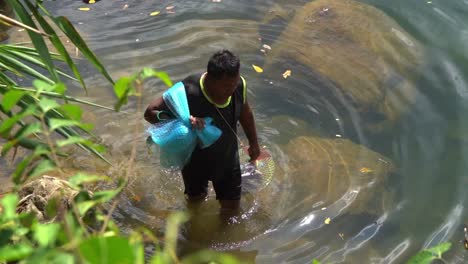 Ein-Thailändischer-Mann-Geht-Mit-Einem-Blauen-Netz-Auf-Der-Schulter-Und-Einem-Fischkäfig-In-Zeitlupe-An-Einem-Fluss-Entlang