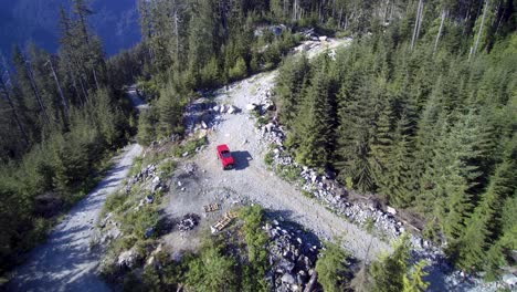 Eine-Aufschlussreiche-Rückansicht-Aus-Der-Luft-Eines-Jeeps,-Der-Auf-Einem-Von-Bäumen-Umgebenen-Nebenstraßenweg-Geparkt-Ist,-Mit-Bergen-Und-Blauem-Himmel-Im-Hintergrund