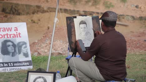 Artista-De-Bocetos-Callejeros-Con-Sombrero-Realiza-Un-Retrato-A-Lápiz-En-Vivo-De-Un-Niño-Pequeño,-Toma-Una-Fotografía-De-Un-Teléfono-Móvil-Y-La-Dibuja-En-Un-Papel-De-Lienzo-Mientras-Se-Sienta-En-Una-Pequeña-Silla-Plegable