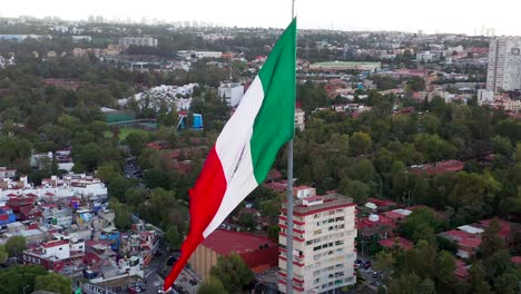 Vuelo-En-Círculo-Por-Una-Gran-Y-Majestuosa-Bandera-Patriótica-Mexicana-Roja,-Blanca-Y-Verde-Ondeando-En-El-Viento-Sobre-El-Centro-De-La-Ciudad-De-México,-Los-Edificios-Y-La-Comunidad-De-San-Jerónimo,-En-Un-Día-Nublado,-Aéreo