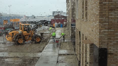 Marco-De-Ventana-Húmedo-Con-Trabajadores-De-La-Construcción-Que-Operan-Maquinaria-Pesada-Para-Colocar-Una-Acera-Frente-A-Casas-Recién-Construidas-En-Un-Día-Lluvioso