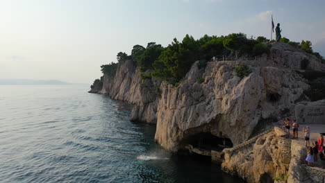 Young-male-backflipping-off-clifftop-into-ocean,-aerial-reveal