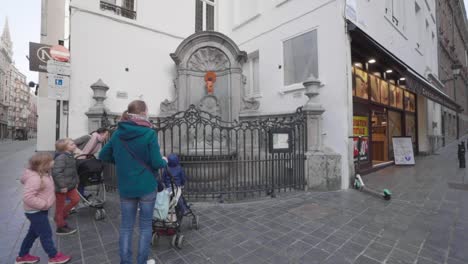 Familia-Con-Mascarilla-Visita-Manneken-Pis-Durante-Covid-19-En-El-Centro-De-Bruselas,-Bélgica---Madres-Empujando-El-Cochecito-De-Bebé-Hacia-La-Escultura-De-La-Fuente-De-Bronce