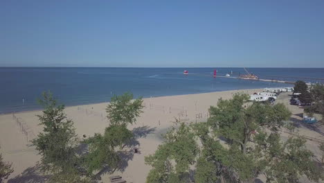 Rising-aerial-view-over-a-sandy-beach-campground-with-playground-and-beach-volleyball-nets-that-is-next-to-a-large-lake-with-a-pier-and-a-lighthouse-under-construction