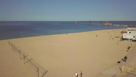 Eine-Sich-Bewegende-Drohne-Schoss-An-Amerikanischen-Flaggen--Und-Beachvolleyballplätzen-Vorbei-über-Einem-Campingplatz-Neben-Einem-Großen-Sandstrand-Mit-Einem-Großen-See-Im-Hintergrund-Und-Einem-Im-Bau-Befindlichen-Pier-Mit-Einem-Leuchtturm