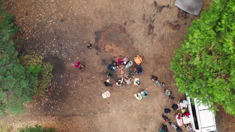 Toma-Aérea-Revelada-De-Jóvenes-Alrededor-De-Una-Fogata-Asando-Perritos-Calientes-Mientras-Acampan-En-El-Bosque