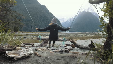 Schwenkaufnahme-Eines-Mädchens,-Das-Auf-Einer-Holzschaukel-Schwingt,-Mit-Schneebedeckten-Gipfeln-Und-Fjorden-Im-Hintergrund