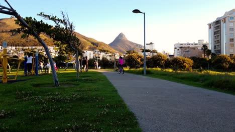 Parque-De-Punto-Verde,-Zona-De-Recreo,-Niños-Jugando---Bicicletas-De-Niñas-Aunque-Fotografiadas-Con-Vistas-A-Sudáfrica,-Ciudad-Del-Cabo-Cabeza-De-Leones-Pico-De-La-Montaña-A-Lo-Largo-Del-Horizonte