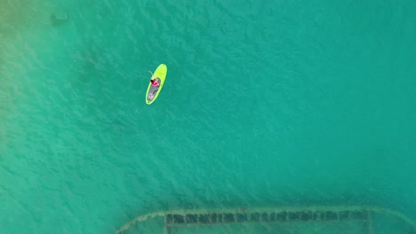 Vista-De-Pájaro-De-Una-Mujer-Remando-De-Pie-Sobre-Un-Naufragio-Oxidado-En-Aguas-Turquesas-Claras-En-Una-Isla-Tropical