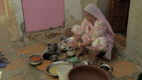 In-this-image,-an-Indian-woman-is-seen-washing-utensils-outside-her-house