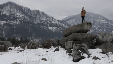 Una-Niña-Se-Para-Sobre-Rocas-Con-Montañas-Nevadas-A-Su-Alrededor