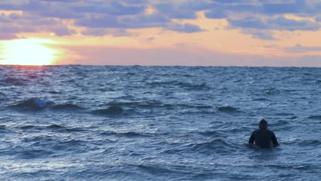 Surfer-on-surfboard-paddling-over-waves-near-the-Baltic-sea-Karosta-beach-at-Liepaja-during-a-beautiful-vibrant-sunset-at-golden-hour,-medium-shot-from-a-distance