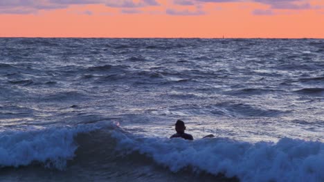 Surfista-En-Tabla-De-Surf-Remando-Sobre-Olas-Cerca-De-La-Playa-De-Karosta-Del-Mar-Báltico-En-Liepaja-Durante-Una-Hermosa-Puesta-De-Sol-Vibrante-A-La-Hora-Dorada,-Plano-Medio-Desde-La-Distancia