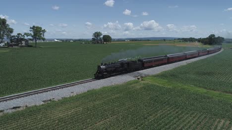 Vista-Aérea-De-Las-Tierras-De-Cultivo-Y-El-Campo-Con-Un-Tren-De-Vapor-Antiguo-Que-Resopla-En-Un-Día-Soleado-De-Verano