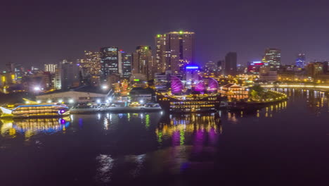 Hyperlapse-Drohnenkamerafahrt,-Die-Die-Uferpromenade-Von-Saigon-Scannt-Und-Die-Wichtigsten-Touristenattraktionen-Und-Die-Wichtige-Infrastruktur-Der-Docks-Und-Des-Finanzzentrums-Bei-Nacht-Mit-Spiegelungen-Im-Saigon-Fluss-Zeigt