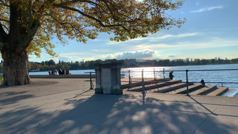 Die-Sonne-Scheint-Auf-Den-See,-Ein-Wunderschöner-Baum-Spendet-Schatten-Auf-Den-Boden-Und-Im-Hintergrund-Sind-Nicht-Wiederzuerkennende-Menschen-Zu-Sehen