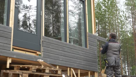 Senior-man-uses-screwdriver-to-tighten-board-to-exterior-wall,-then-uses-a-hand-held-cirkular-saw-to-cut-the-board-into-the-right-length,-static-handheld-wide-shot