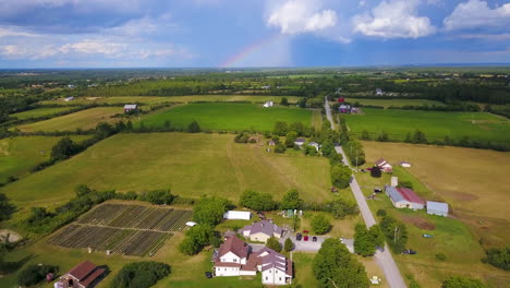 Elevación-Aérea-Sobre-Una-Granja-Con-Un-Arco-Iris-Y-Nubes-De-Tormenta-En-La-Distancia