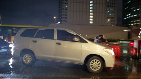 Outdoor-evening-scene-showing-vehicular-traffic-in-a-drizzle-around-a-commercial-district-within-Quezon-City-in-Metro-Manila,-Philippines