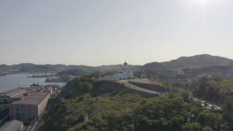 Toma-Aérea-De-Pivote-Lento-De-Una-Iglesia-Ortodoxa-Con-Techo-Azul-Y-Cúpulas-Doradas,-Ubicada-En-La-Cima-De-La-Colina-Con-El-Puerto-Y-El-Edificio-De-La-Ciudad-En-El-Fondo,-En-Un-Día-Brillante,-Claro-Y-Soleado