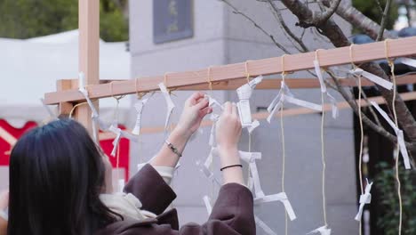 Japanese-girl-hanging-paper-note-on-rope-attached-from-wooden-frame-at-Yasaka-Shrine-in-Kyoto,-Japan