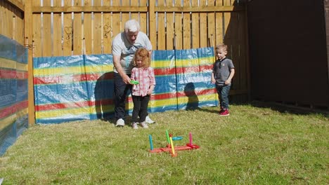 Abuelo-Jugando-Un-Juego-Con-Su-Nieta-En-El-Jardín-Trasero-Mientras-Un-Niño-Pequeño-Los-Observa-Durante-El-Verano