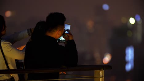 Zwei-Asiatische-Frauen-Und-Ein-Mann-Fotografieren-Mit-Ihren-Mobiltelefonen-Die-Nächtliche-Skyline-Der-Stadt,-Nahaufnahme-Aus-Der-Hand