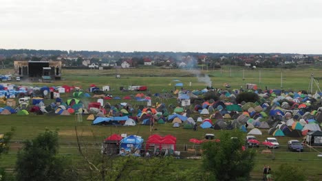 Beim-Flug-über-Bäume-Kommen-Bunte-Zelte-Und-Eine-Bühne-Auf-Einem-Feld-Bei-Einem-Musikfestival-Zum-Vorschein,-Mit-Der-Stadt-Im-Hintergrund