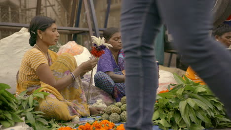 Women-sitting-around-a-pile-of-leaves,-fruits-and-flowers-arranging-them-into-bouquets-as-people-walk-by