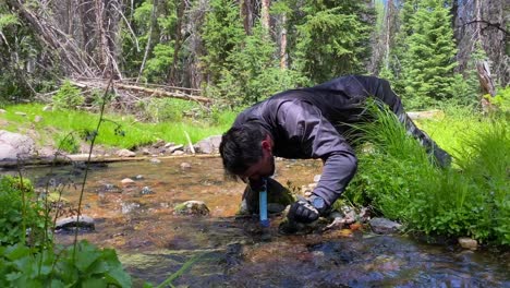 Hombre-Vestido-Con-Equipo-De-Bicicleta-De-Tierra-Bebiendo-Agua-De-Un-Arroyo-Salvaje-Usando-Un-Sistema-De-Filtro-De-Agua-Limpia-De-Paja-De-Vida