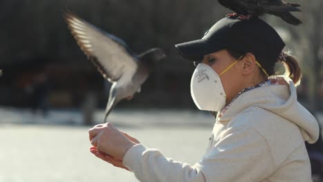 Mujer-Joven-Con-Mascarilla-Y-Sombrero-Divirtiéndose-Con-Palomas-Del-Parque-En-Brazos-Y-Cabeza-En-Un-Día-Soleado-Durante-La-Pandemia-Del-Virus-Covid-19-60fps