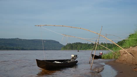 Lange-Boote-Mit-Motoren-Vertäut-Und-Hüpfen-Auf-Dem-Mekong-Fluss,-Während-Ein-Bootsmann-Sein-Langboot-In-Richtung-Der-Linken-Seite-Des-Bildes-Beschleunigt,-Mit-Berghintergrund-Und-Schönem-Blauen-Himmel