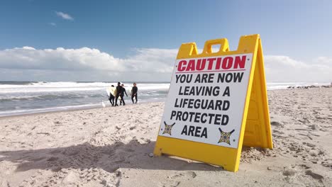 Los-Surfistas-Caminan-Por-El-Océano-Detrás-De-La-Señal-De-Precaución-De-La-Playa-En-El-Soleado-Día-De-Florida