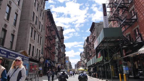 Two-Men-in-Wheelchairs-Move-Down-NYC-Street,-Camera-Pan-Up,-Bird-Flying-Sun-Flare,-Wide-Shot