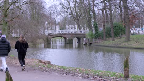 Two-friends-walking-through-a-park-together,-along-the-canal-in-Utrecht,-Netherlands,-with-a-beautiful-bridge-in-the-background