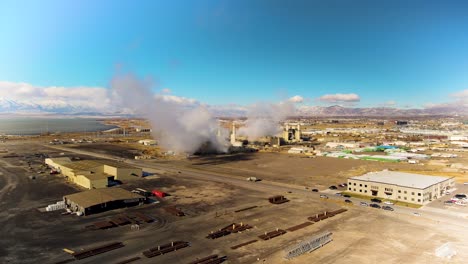 Un-Complejo-Industrial-Emite-Humo-Y-Vapor-Al-Aire-Con-Una-Vista-Aérea-De-Un-Lago-Y-Montañas-Nevadas