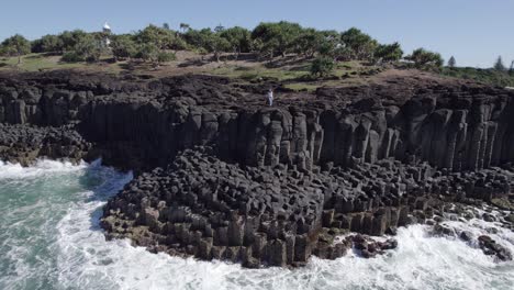 Female-Tourist-Standing-On-Basalt-Columns-Of-Fingal-Head-Causeway-With-Sea-Waves-Crashing-In-NSW,-Australia