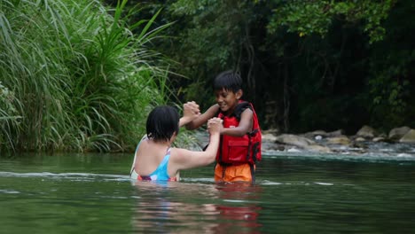 Niño-Indio-Con-Chaleco-Salvavidas-Rojo-Juguetonamente-Luchando-A-Mano-Con-Una-Mujer-Asiática-En-El-Río