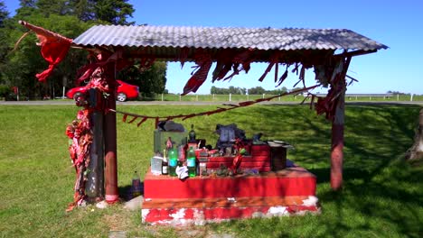 Un-Auto-Rojo-Pasa-Detrás-De-Un-Santuario-En-Honor-Al-Gauchito-Gil-Cerca-De-Una-Ruta-Provincial