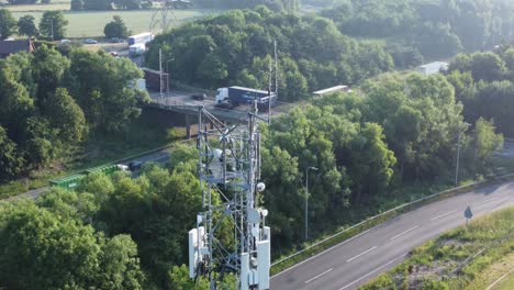 Antena-De-Torre-De-Radiodifusión-5g-En-El-Campo-Británico-Con-Vehículos-Que-Viajan-En-La-Vista-Aérea-Inclinada-Hacia-Arriba-Del-Fondo-De-La-Carretera