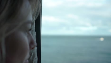 Slowmo---Junge-Schöne-Frau-Mit-Blonden-Haaren,-Die-Aus-Dem-Fenster-Mit-Blick-Auf-Das-Meer-Schaut---Nahaufnahme