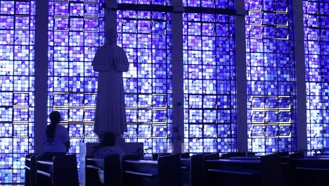 Tiro-Largo-De-Dos-Personas-Rezando-Frente-A-Una-Escultura-En-La-Iglesia-Dom-Bosco-Iluminada-Desde-Las-Ventanas-Azules