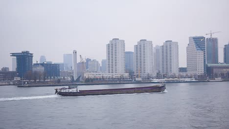 Großes-Frachtschiff,-Das-An-Einem-Regnerischen,-Bewölkten-Tag-Durch-Die-Stadt-Rotterdam-Fährt,-Mit-Teilweise-Im-Bau-Befindlichen-Wolkenkratzern-Des-Finanzviertels-Im-Hintergrund