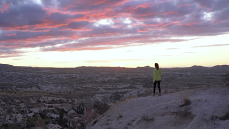 Mujer-Caminando-Hacia-El-Borde-De-Los-Acantilados-Para-Admirar-La-Puesta-De-Sol-En-El-Valle-Rojo-En-Goreme,-Turquía